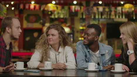 Portrait-of-cheerful-young-friends-looking-at-smart-phone-while-sitting-in-cafe.-Mixed-race-people-sitting-at-a-table-in-restaurant-using-mobile-phone